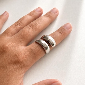 May include: A polished silver-toned ring featuring a thick, smooth, and rounded band design. The ring has a high-shine metallic finish with a curved, chunky silhouette that wraps partially around the finger, creating a bold and reflective appearance.
