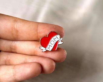 Könnte beinhalten: Rote herzförmige Emaille-Anstecknadel mit weißem Banner mit der Aufschrift „MOM“. Die Anstecknadel hat eine schwarze Umrandung. Ein schönes Geschenk zum Muttertag oder zu jedem Anlass.