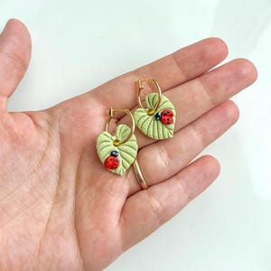 Pode incluir: Brincos de argola dourados com pingentes em forma de folha. Os pingentes verde claro têm veias detalhadas e uma joaninha vermelha e preta. Os brincos são segurados em uma mão.