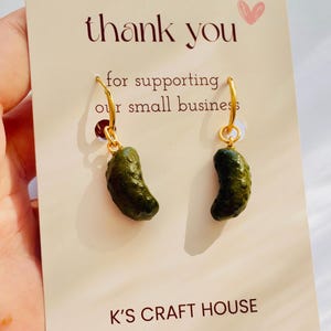 May include: A pair of gold-toned hoop earrings featuring small, textured, dark green pickle charms. The earrings are attached to a cream-colored display card with the text thank you for supporting our small business and K'S CRAFT HOUSE.