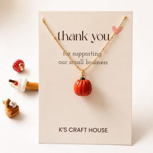 Può includere: Una collana a catena dorata con un piccolo ciondolo a forma di zucca arancione su un cartoncino color crema con la scritta thank you for supporting our small business e K'S CRAFT HOUSE. Il cartoncino presenta anche un piccolo cuore rosa.