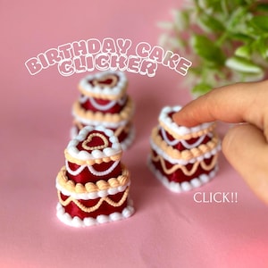 Puede incluir: Miniaturas de clickers de pastel de cumpleaños rojos, blancos y beige. Los pasteles tienen una capa superior en forma de corazón y están decorados con glaseado blanco y adornos beige. El texto "BIRTHDAY CAKE CLICKER" está arqueado sobre los pasteles, y "CLICK!!" está escrito debajo.