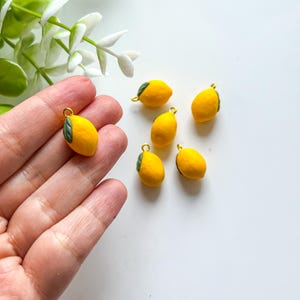 Könnte beinhalten: Kleine gelbe Zitronen-Anhänger mit grünen Blättern und goldfarbenen Schlaufen. Die Anhänger sind detailliert und haben eine strukturierte Oberfläche. Mehrere Anhänger liegen auf einer weißen Oberfläche.