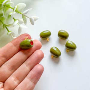 Può includere: Ciondoli a forma di oliva, con esterno verde e centro rosso. Ogni ciondolo ha un anello dorato per l'attacco. I ciondoli sono esposti su uno sfondo bianco, suggerendo un tema artigianale o di gioielleria.