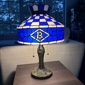 May include: A stained glass table lamp with a blue and white chequered shade. The shade features a blue diamond with a white "B" in the centre. The lamp has a bronze base.