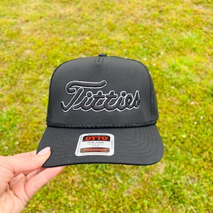 May include: Black trucker hat with the word "Titties" embroidered in white script. The hat has a mesh back and a curved brim. The hat is held by a person in front of a green grassy background.