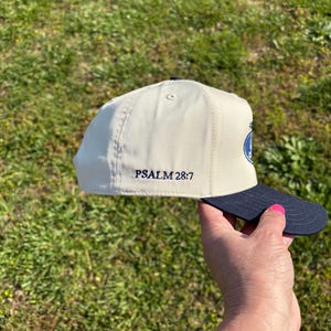 May include: A cream-colored baseball cap with a dark navy blue brim. The side of the cap features the embroidered text PSALM 28:7 in dark navy blue thread. The cap has a structured crown with a button on top and visible stitching along the panels.