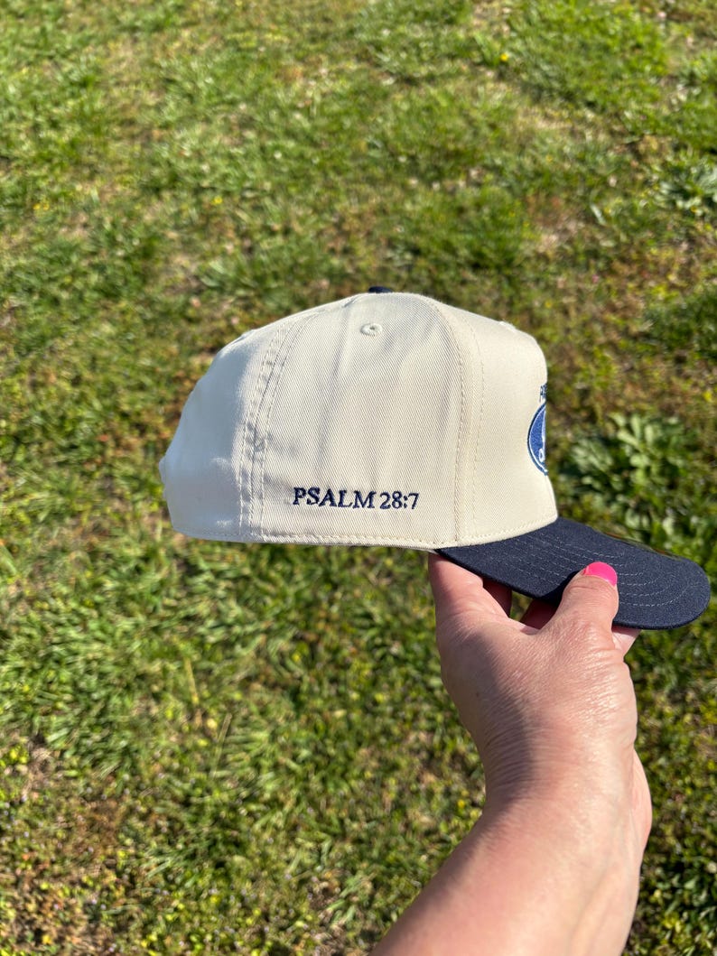May include: A cream-colored baseball cap with a dark navy blue brim. The side of the cap features the embroidered text PSALM 28:7 in dark navy blue thread. The cap has a structured crown with a button on top and visible stitching along the panels.