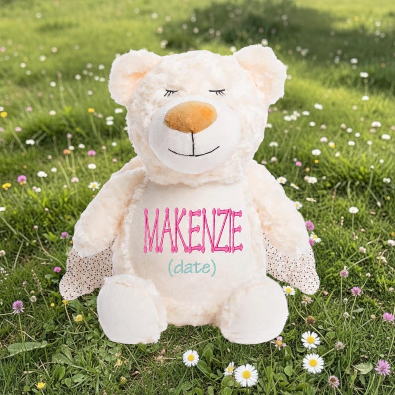 Op de afbeelding: Een cr&egrave;mekleurige teddybeer met de naam "MAKENZIE" geborduurd in roze script. De beer heeft een lichtbruine neus, gesloten ogen en gestippelde accenten op zijn poten. De tekst "(date)" staat onder de naam in teal.