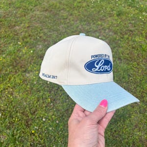 May include: A cream-colored baseball cap with a light blue brim. The front features a dark blue oval emblem with the text "POWERED BY THE Lord" in a script font. The side of the cap includes the embroidered text "PSALM 28:7" in dark blue thread.
