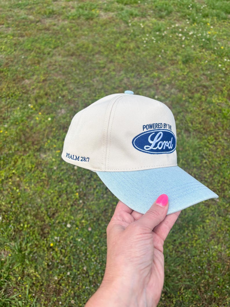 May include: A cream-colored baseball cap with a light blue brim. The front features a dark blue oval emblem with the text "POWERED BY THE Lord" in a script font. The side of the cap includes the embroidered text "PSALM 28:7" in dark blue thread.
