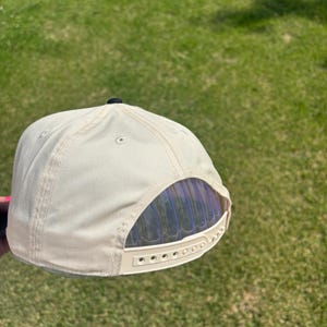 May include: A cream-colored baseball cap featuring a structured six-panel design with a small black button at the top. The back of the cap includes an adjustable plastic snapback closure with the brand name Otto embossed on the strap.