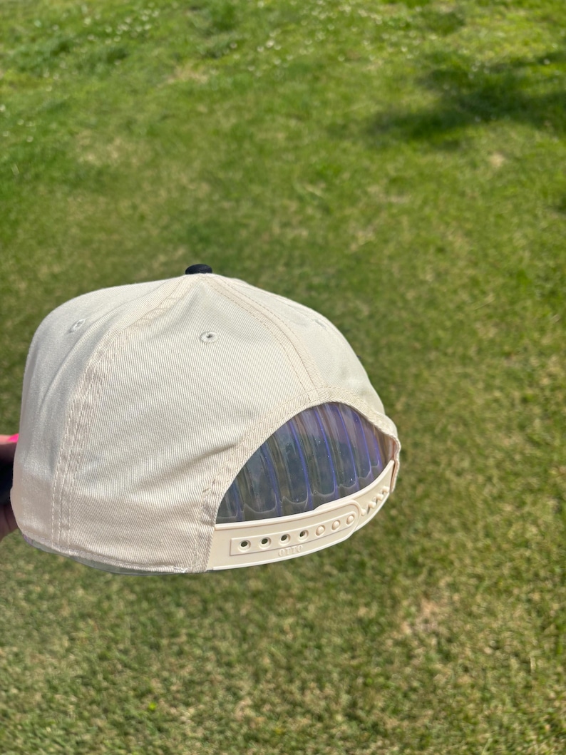 May include: A cream-colored baseball cap featuring a structured six-panel design with a small black button at the top. The back of the cap includes an adjustable plastic snapback closure with the brand name Otto embossed on the strap.