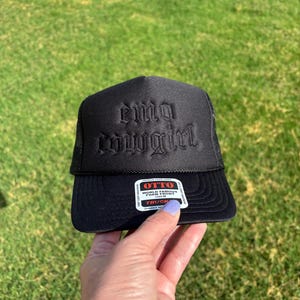 May include: A black trucker hat featuring the embroidered text "emo cowgirl" in a gothic font on the front foam panel. The hat includes a black mesh back and a sticker on the brim that reads "OTTO WORLD FAMOUS FOAM FRONT OSFM TRUCK".
