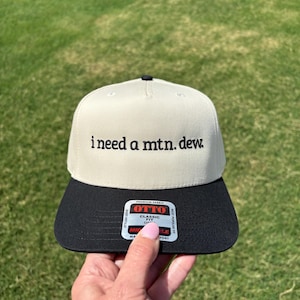 May include: A beige baseball cap with a black brim featuring the embroidered black text "i need a mtn. dew." on the front panel. A rectangular sticker with the brand name "OTTO" and "CLASSIC FIT" is attached to the center of the black brim.