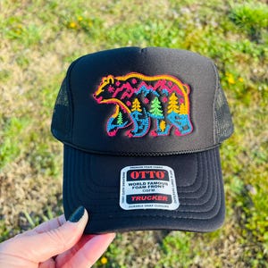 May include: Black trucker hat with a colorful bear design. The bear is outlined in yellow and filled with pink, blue, and green trees. The hat has a mesh back and a curved brim. The hat has the text "OTTO WORLD FAMOUS FOAM FRONT OSFM TRUCKER DURABLE SNAP CLOSURE".