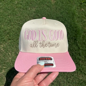 May include: A cream-colored baseball cap with a pink brim featuring the embroidered text "GOD IS GOOD" in pink and "all the time" in brown. A rectangular sticker on the brim reads "OTTO CLASSIC FIT PREMIUM FABRIC PROFILE".