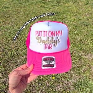 May include: A pink trucker hat with a white front panel. The text "PUT IT ON MY Daddy's TAB" is embroidered in gold. The hat has a pink brim and mesh back. The text "Choose Any Hat Style and Color" is also visible.