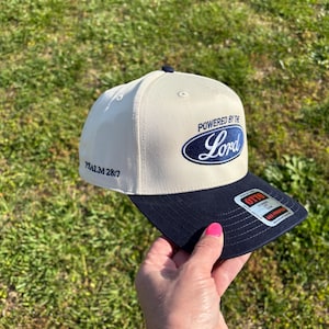 May include: A cream-colored baseball cap with a navy blue brim and a navy blue oval logo on the front featuring the text "POWERED BY THE LORD" in white script. The side of the cap displays the text "PSALM 28:7" in navy blue, and a sticker is on the brim.