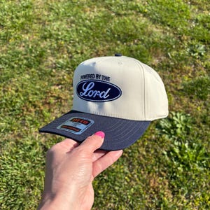 May include: A beige baseball cap with a navy blue brim and a navy blue oval patch on the front. The patch features the text "POWERED BY THE" in navy blue block letters above the word "Lord" in a white script font. An Otto brand sticker is on the brim.