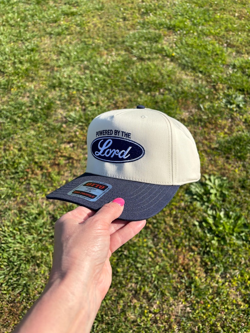 May include: A beige baseball cap with a navy blue brim and a navy blue oval patch on the front. The patch features the text "POWERED BY THE" in navy blue block letters above the word "Lord" in a white script font. An Otto brand sticker is on the brim.