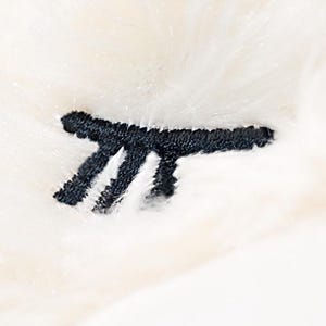 Op de afbeelding: Close-up van een zacht, cr&egrave;mekleurig pluche materiaal met een zwart geborduurd ontwerp. Het ontwerp lijkt een gestileerde letter of symbool te zijn, wat een vleugje detail toevoegt aan de donzige textuur.