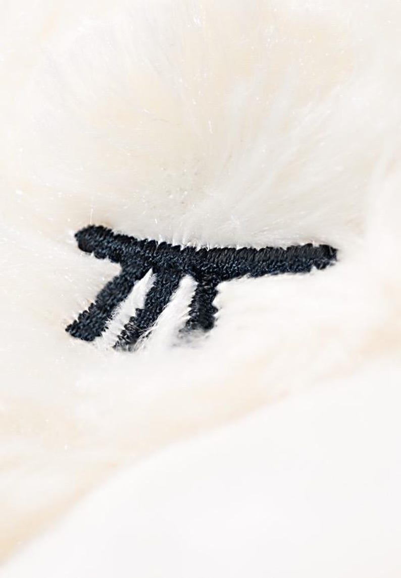 Op de afbeelding: Close-up van een zacht, cr&egrave;mekleurig pluche materiaal met een zwart geborduurd ontwerp. Het ontwerp lijkt een gestileerde letter of symbool te zijn, wat een vleugje detail toevoegt aan de donzige textuur.