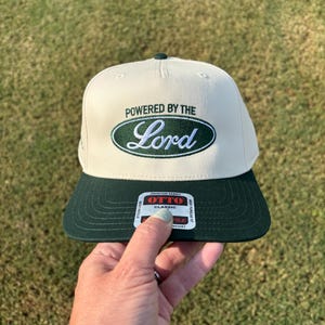 May include: A cream-colored baseball cap with a dark green brim and a dark green oval patch on the front. The patch features the embroidered text "POWERED BY THE" in dark green and the word "Lord" in white script. An Otto Classic sticker is on the brim.
