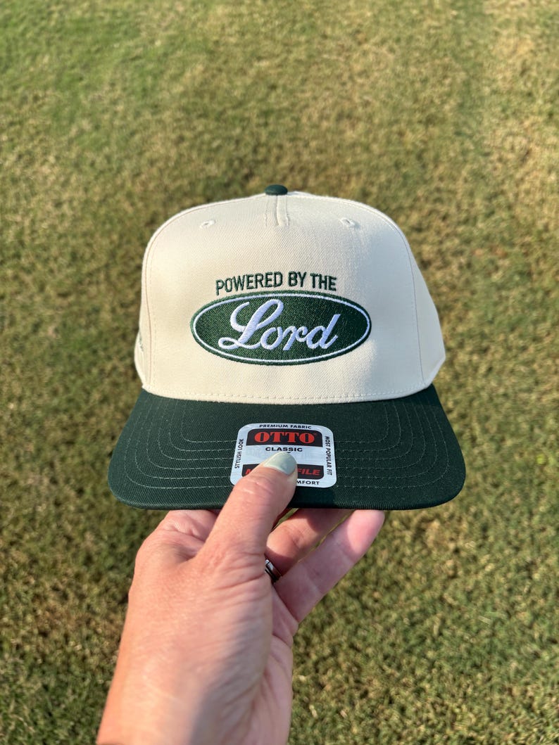 May include: A cream-colored baseball cap with a dark green brim and a dark green oval patch on the front. The patch features the embroidered text "POWERED BY THE" in dark green and the word "Lord" in white script. An Otto Classic sticker is on the brim.