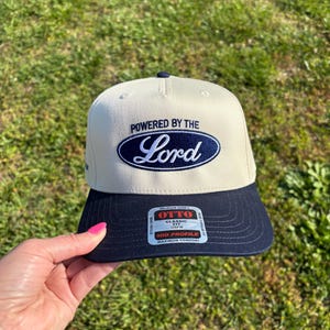 May include: A cream-colored baseball cap with a navy blue brim. The front features an embroidered navy blue oval logo with the text "POWERED BY THE Lord" in white and blue. A rectangular sticker on the brim reads "OTTO CLASSIC FIT OSFM".