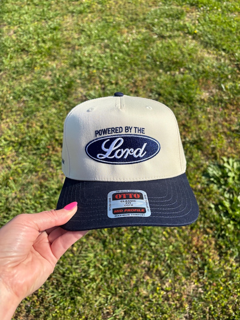 May include: A cream-colored baseball cap with a navy blue brim. The front features an embroidered navy blue oval logo with the text "POWERED BY THE Lord" in white and blue. A rectangular sticker on the brim reads "OTTO CLASSIC FIT OSFM".
