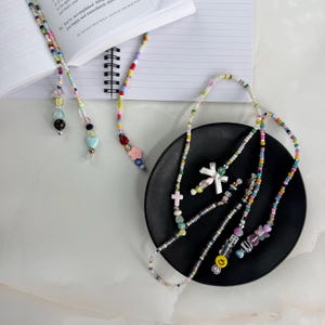 May include: Colorful beaded bookmarks with various charms, including hearts, crosses, and flowers. The bookmarks are displayed on a black plate and next to an open book. The beads are in a variety of colors, including pink, blue, yellow, and green.