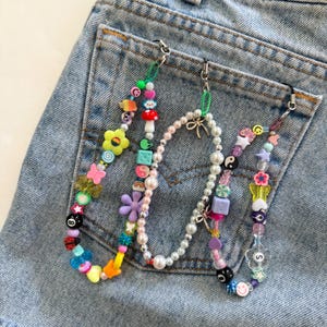 May include: Three beaded phone charms with colorful beads, including flowers, smiley faces, and stars. The charms are attached to a denim pocket. The beads are in various colors, including pink, yellow, blue, and green.