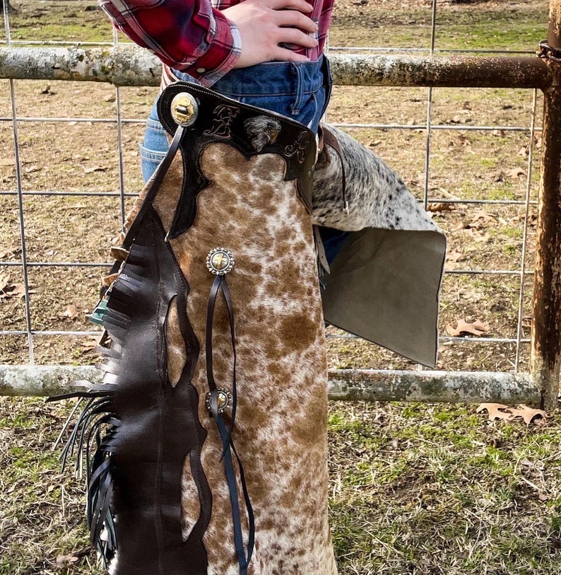 May include: Cowhide chaps with brown and white spotted pattern. The chaps have dark brown leather accents, decorative silver-colored conchos, and fringe along the side. The chaps are designed for riding and have a western theme.