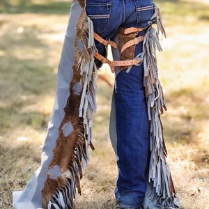 May include: A pair of denim jeans with brown leather chaps. The chaps have a light blue and brown color scheme with fringe detailing. The chaps are secured with leather straps and buckles. The person is standing on dry grass.