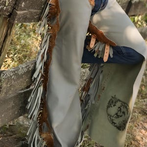 May include: Gray leather chaps with brown cowhide accents and fringe detailing. The chaps feature leather straps and a decorative emblem. The chaps are designed for riding and outdoor activities.