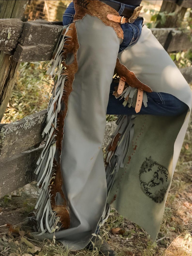 May include: Gray leather chaps with brown cowhide accents and fringe detailing. The chaps feature leather straps and a decorative emblem. The chaps are designed for riding and outdoor activities.