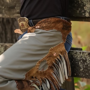 May include: Close-up of a pair of cowboy chaps in shades of brown and gray. The chaps feature a fringed design and are worn over blue jeans. A decorative belt buckle is visible.