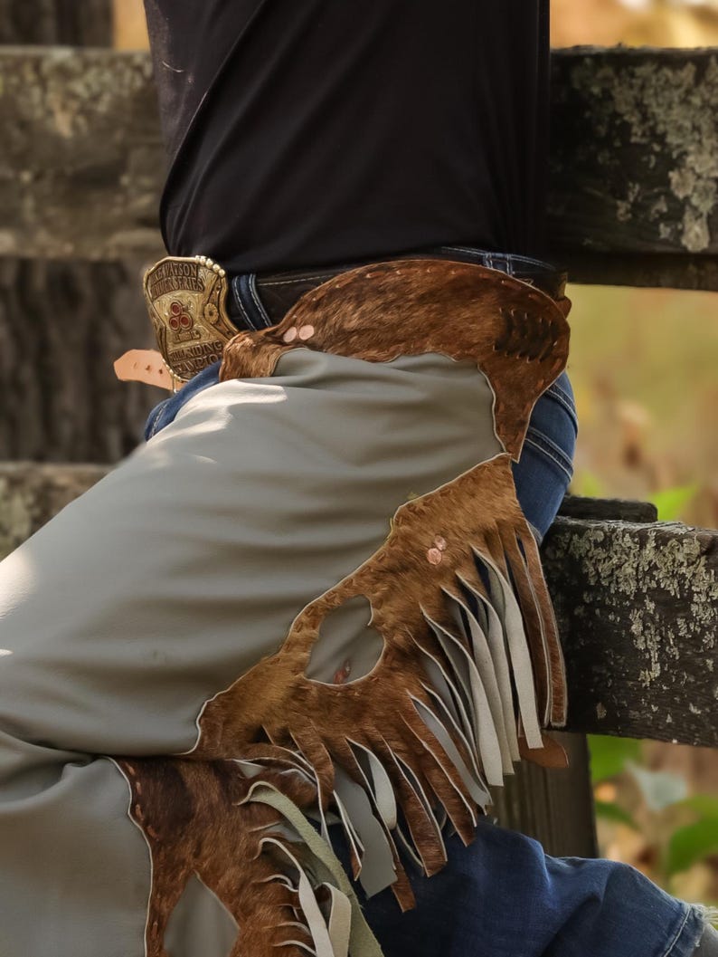 May include: Close-up of a pair of cowboy chaps in shades of brown and gray. The chaps feature a fringed design and are worn over blue jeans. A decorative belt buckle is visible.