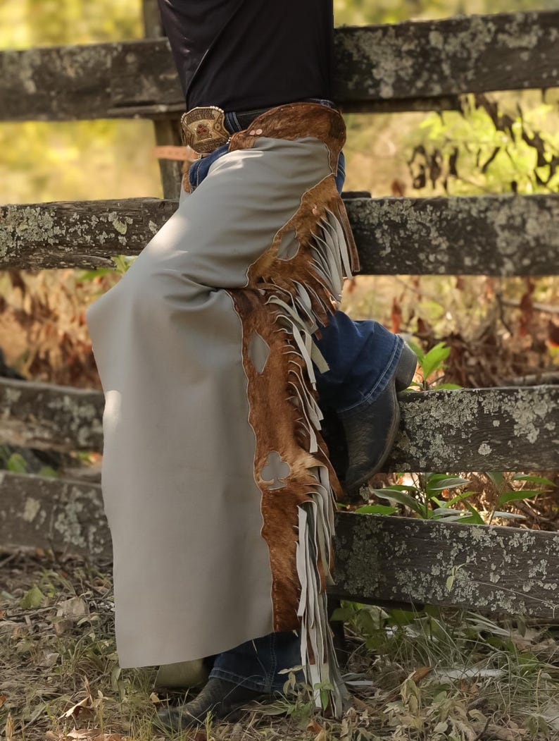 May include: Gray leather chaps with brown fur trim and white fringe. The chaps are worn over blue jeans and black cowboy boots. A decorative belt buckle is visible. The chaps are leaning against a weathered wooden fence.