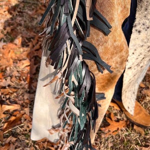 May include: Close-up of a pair of leather chaps with a mix of brown, black, and green fringe. The chaps have a light tan suede panel and a brown and white cowhide pattern. The chaps are worn with brown boots.