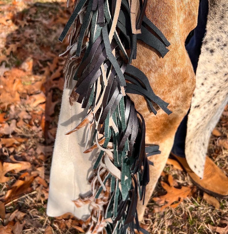 May include: Close-up of a pair of leather chaps with a mix of brown, black, and green fringe. The chaps have a light tan suede panel and a brown and white cowhide pattern. The chaps are worn with brown boots.
