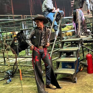 May include: A cowboy in a plaid shirt, leather vest, and chaps with red accents stands near a metal staircase. Two other cowboys are visible in the background, one seated and another standing. The scene is set in an arena.