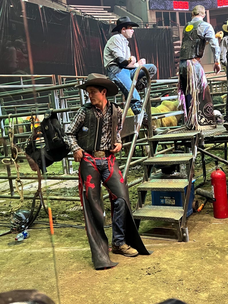 May include: A cowboy in a plaid shirt, leather vest, and chaps with red accents stands near a metal staircase. Two other cowboys are visible in the background, one seated and another standing. The scene is set in an arena.
