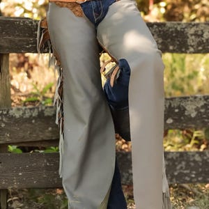 May include: Gray leather chaps with fringe detail, worn over blue jeans. A brown leather belt with a decorative buckle and a brown leather accent is visible. The chaps are knee-length and appear to be made of a durable material.