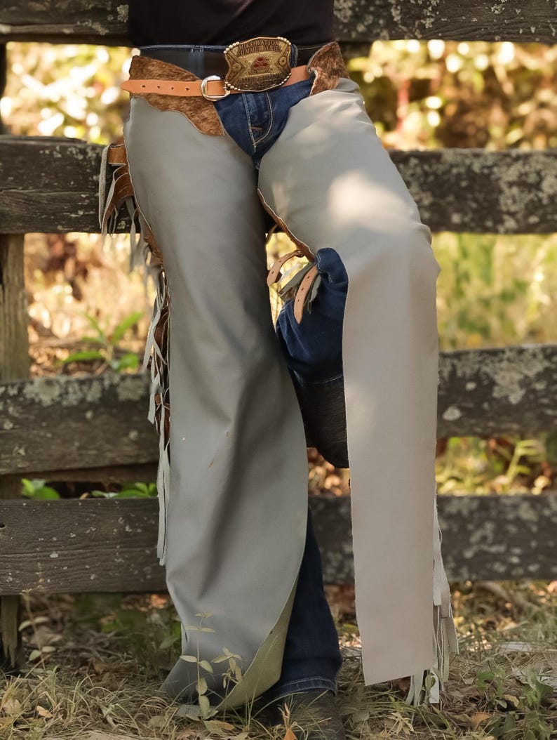 May include: Gray leather chaps with fringe detail, worn over blue jeans. A brown leather belt with a decorative buckle and a brown leather accent is visible. The chaps are knee-length and appear to be made of a durable material.
