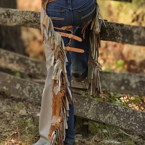 May include: Leather chaps with fringe and buckle details. The chaps are tan and brown, with a distressed look. The fringe hangs down the sides of the legs. The chaps are worn with a brown belt and blue jeans.
