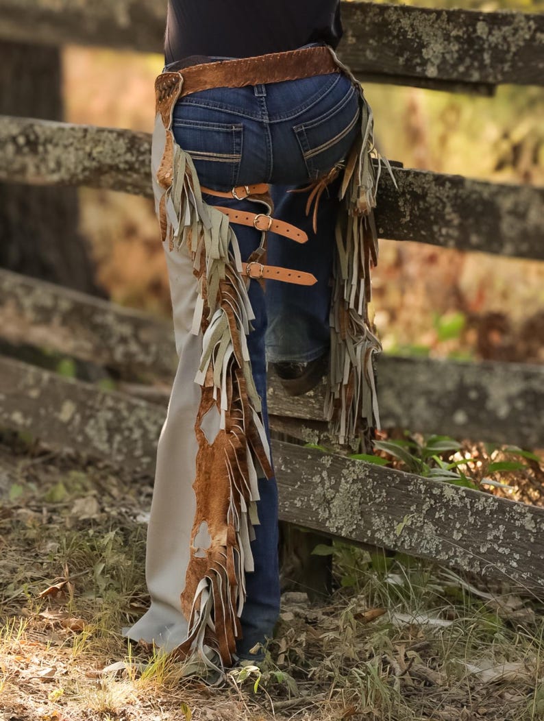 May include: Leather chaps with fringe and buckle details. The chaps are tan and brown, with a distressed look. The fringe hangs down the sides of the legs. The chaps are worn with a brown belt and blue jeans.