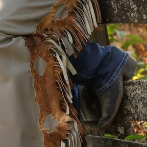 May include: Brown and tan leather chaps with fringe and a club-shaped cutout. The chaps are paired with blue jeans and black cowboy boots, resting on a weathered wooden fence. The chaps have a light tan lining.