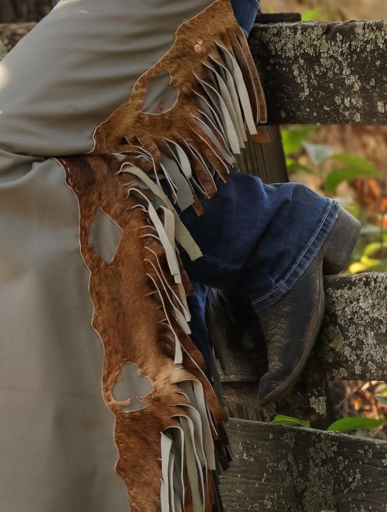 May include: Brown and tan leather chaps with fringe and a club-shaped cutout. The chaps are paired with blue jeans and black cowboy boots, resting on a weathered wooden fence. The chaps have a light tan lining.